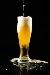 Vertical image of lager beer dripping into overflowing pint glass on black, with copy space. Drinking alcohol, refreshment and lager day celebration concept.