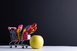 Close up of miniature shopping trolley school materials, apple and copy space on grey background. Back to school, shopping, school materials, learning, school and education concept.
