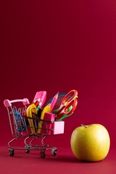 Close up of miniature shopping trolley with school materials, apple and copy space on red background. Back to school, shopping, school materials, learning, school and education concept.