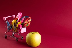 Close up of miniature shopping trolley with school materials, apple and copy space on red background. Back to school, shopping, school materials, learning, school and education concept.