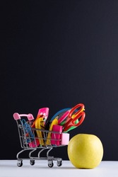 Close up of miniature shopping trolley school materials, apple and copy space on black background. Back to school, shopping, school materials, learning, school and education concept.