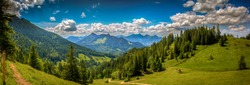 Beautiful alpine landscape with green hills and conifer forest, clouds in blue sky. Picturesque panoramic view of nature