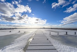 A pier in the sea by the entrance to the port with frozen sea and snow, in the background blue sky, sun and white clouds, ultra wide angle of view.
