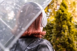 Autumn. Lonely sad redhead woman in a headphones walking in a park, garden. View through wet transparent umbrella with rain drops. Rainy day landscape. 