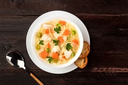 An overhead photo of a plate of chicken, vegetables, and noodles soup, shot from above on a dark rustic texture with slices of bread and a spoon, and a place for text