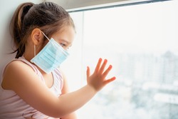 Little european girl wearing mask for protect pm2.5 and Covid-19. Sick child Little girls look at the window with longing on the street . Copy  space. sadness child at home in isolation           
