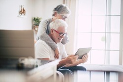 couple of seniors smiling and looking at the same tablet hugged on the sofa - indoor, at home concept - caucasians mature and retired man and woman using technology - lockdown and quarantine lifestyle