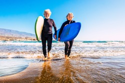 two old and mature people having fun and enjoying their vacations outdoors at the beach wearing wetsuits and holding a surfboard to go surfing in the water with waves - active senior smiling 