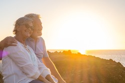 couple of two cute seniors together enjoying summer and having fun at the beach looking at the sea or ocean with sunset - mature people having a good lifestyle