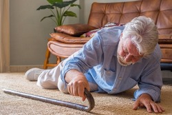 Elder senior man lying on floor after falling down with wooden walking stick beside couch on rug in living room at home. Old man suffering with pain and struggling to get up after falling down at home