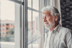 Happy thoughtful older 70s man looking out of window away with hope, thinking of good health, retirement, insurance benefits, dreaming of future. Elderly pensioner waiting meeting with family