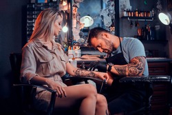 Dilligent focused tattoo artist is creating new tattoo on young woman's hand at tatoo studio.