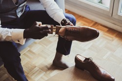 Posh elegant groom is dressing up his leather brown shoes while sitting on sofa.