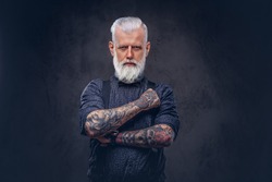 Brutal and fashionable grandpa with gray hairs and tattoos poses in dark background looking at camera.