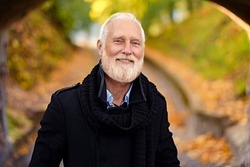 Portrait of joyful elderly man with long beard looking at camera in autum forest under arch.