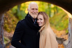 Shot of glad elderly man embracing his young girlfriend in autum forest in daytime.