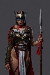 Portrait of woman warrior with tattooed body and spear dressed in light armor staring at camera.