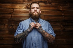 Brutal male with beard and tatooes buttons up his t-shirt