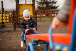 Cute little boy dressed in a warm jacket and hat riding on the swing with parents
