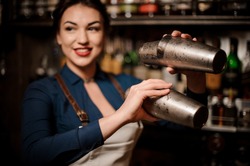 Attractive bartender girl in the white apron holding in her hands two steel cocktail shakers at the bar counter