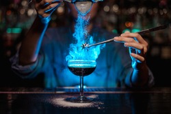 Bartender girl adding to a brown cocktail and pour on a flamed badian on tweezers a powdered sugar on the bar counter in the blue light.
