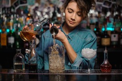 Female bartender pouring to the measuring glass cup with ice cubes an alcoholic drink from steel jigger on the bar counter on the blurred background