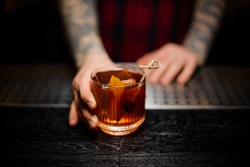 Bartender serving a glass of a Old Fashioned cocktail with orange zest on the bar counter on the blurred background
