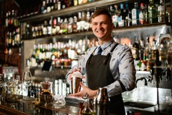 young smiling bartender in black apron preparing to make cocktail. Shelves with bottles of alcohol in background.