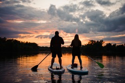 rear view on couple of people standing on sup boards with oars in their hands on the river at sunset