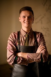 young smiling man bartender in shirt and leather brown apron stands with arms crossed on his chest. Man portrait