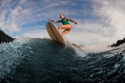 Active female wakesurfer jumping on a wake board on splashing river wave against the cloudy blue sky