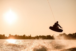 Great view of dark silhouette of active male rider holding rope and making extreme jump on wakeboard over splashing water. Wakeboarding and water sports activity.
