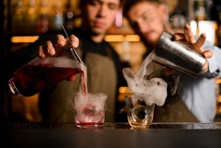 Two glasses stands on bar and two young bartenders fills them with smoke and alcohol at the same time