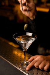 focus on green berry olive over transparent martini glass with cocktail on bar counter. Hand of a male bartender holds a glass.
