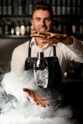 wonderful view of the glassy shaker in the air and the hands of male bartender near it. Blurred bartender and shelves with bottles in the background