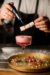 bartender holds skewers with marshmallows above glass with foam and syrup cocktail and burns it using flomber. Glass with cocktail stands on the golden table decorated with raspberries and flovers
