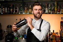Professional bartender in white shirt and black apron holds in hands steel shaker. Blurred bottles on shelves in the background