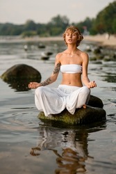 peaceful woman practices yoga position doing meditation in tranquil outdoor sitting on stone in water. Mental wellbeing healthy lifestyle.