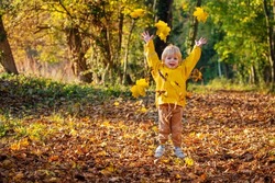Happy little boy in yellow bright coat throwing leaves in sunny autumn forest and jumping joyfully