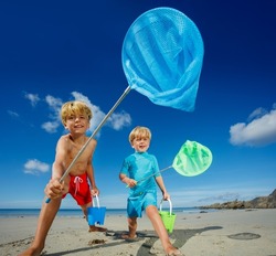 Low angle portrait of two happy blond boys, brothers stand with butterfly nets lean catching critters on the sand ocean beach during tide