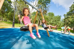Two beautiful little girls swinging on swings on playground with smile on sunny summer day