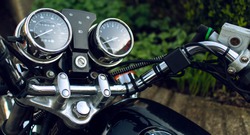The dashboard of a black and silver motorbike. Details of the speed dial and gear dial shown, as well as handles and breaks. There is greenery in the background. Motorbike parked.