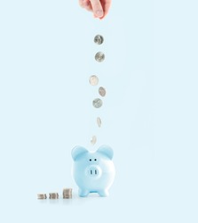Savings concept. Female hand putting coin into piggy bank on pastel blue background