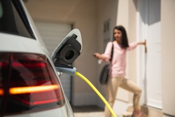 Close up of a electric car charger with female silhouette in the background, entering the home door and locking car