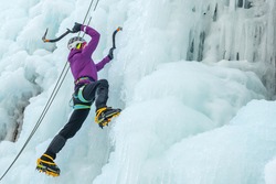 Alpinist woman with ice climbing equipment, axe and climbing ropes, hiking at a frozen waterfall