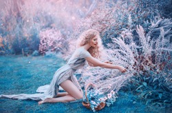 field mermaid collects herbs and flowers in small basket. slender forest nymph sits on her knees in long light dress with open bare legs and back, glare of bright sun in magical purple and blue glade