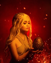 portrait fantasy young beautiful blond woman holds in her hands large unusual dragon egg. Background red flaming fire smoke glow sparks. Studio creative photography Daenerys Targaryen white hair wig 