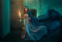 lady Queen woman medieval royal dress run escapes from Gothic night castle. Blue silk dress, cloak train plume waving motion. Holds in hands old candlestick burning candles. Background old retro room