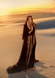 woman in black long dress stands in desert. Luxurious clothes, gold veil accessories hide face. Oriental beauty fashion model. Sand dunes background yellow orange sunset. abaya dress, hijab headscarf
