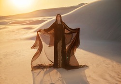A mysterious woman in a black long dress stands in the desert. Luxurious clothes gold accessories hide the face. Oriental beauty fashion model. Sand dunes background orange sunset. hands raised to sky